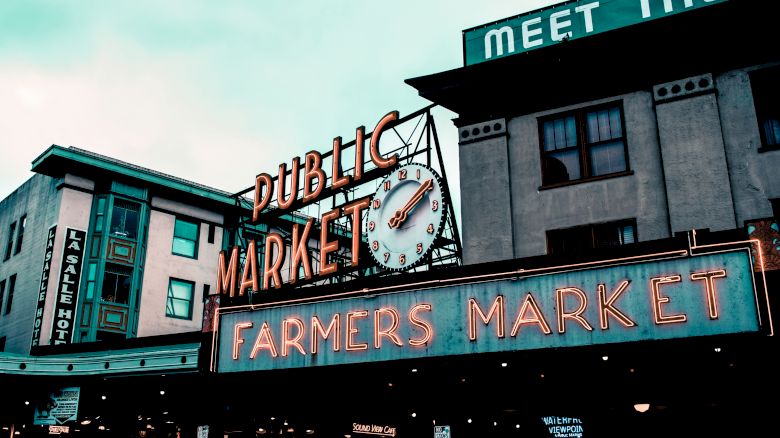 This image shows the entrance to a public market with neon signs reading "PUBLIC MARKET" and "FARMERS MARKET" on a cloudy day, with city buildings around.