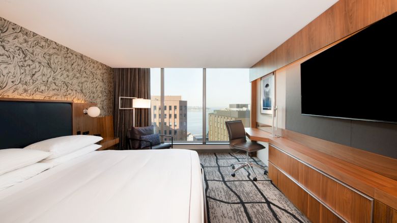 The image shows a modern hotel room with a large bed, desk, chair, flat-screen TV, and a view of city buildings through floor-to-ceiling windows.