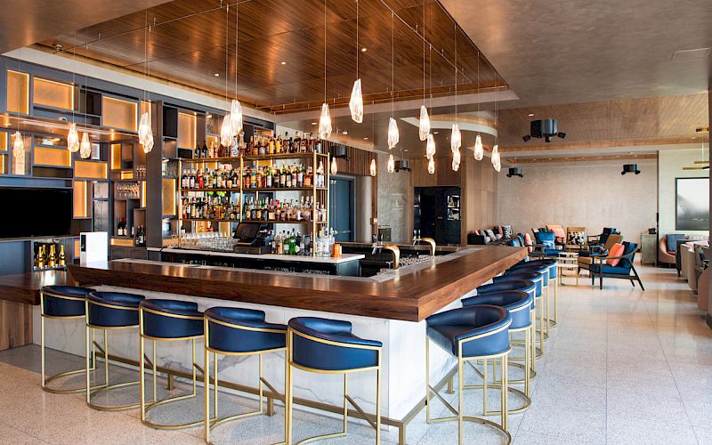 This image shows a modern bar area with a central bar counter, blue bar stools, hanging pendant lights, liquor bottles, and seating in the background.