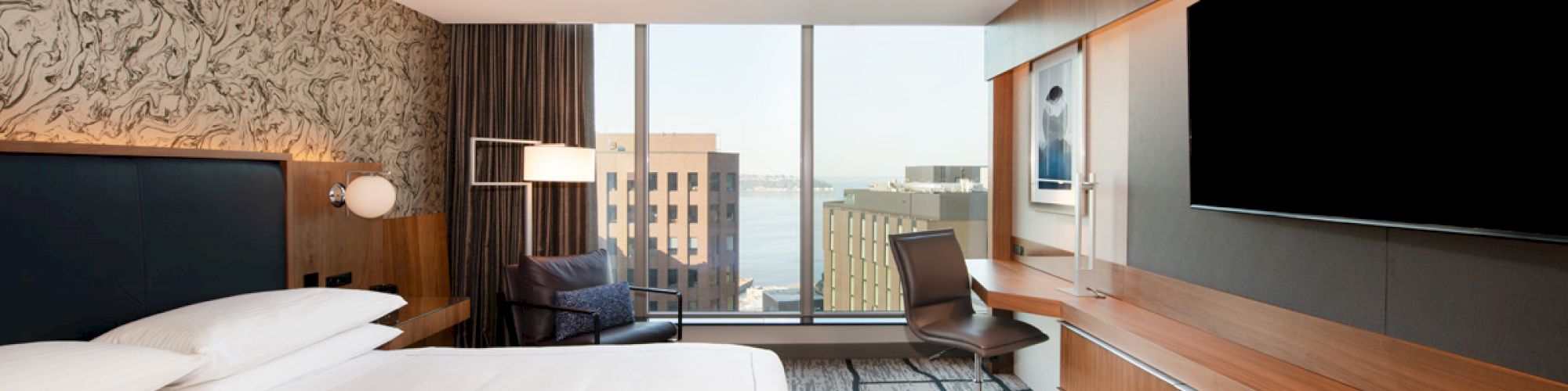 The image shows a modern hotel room with a large bed, desk, chair, television, and a window with a cityscape view.