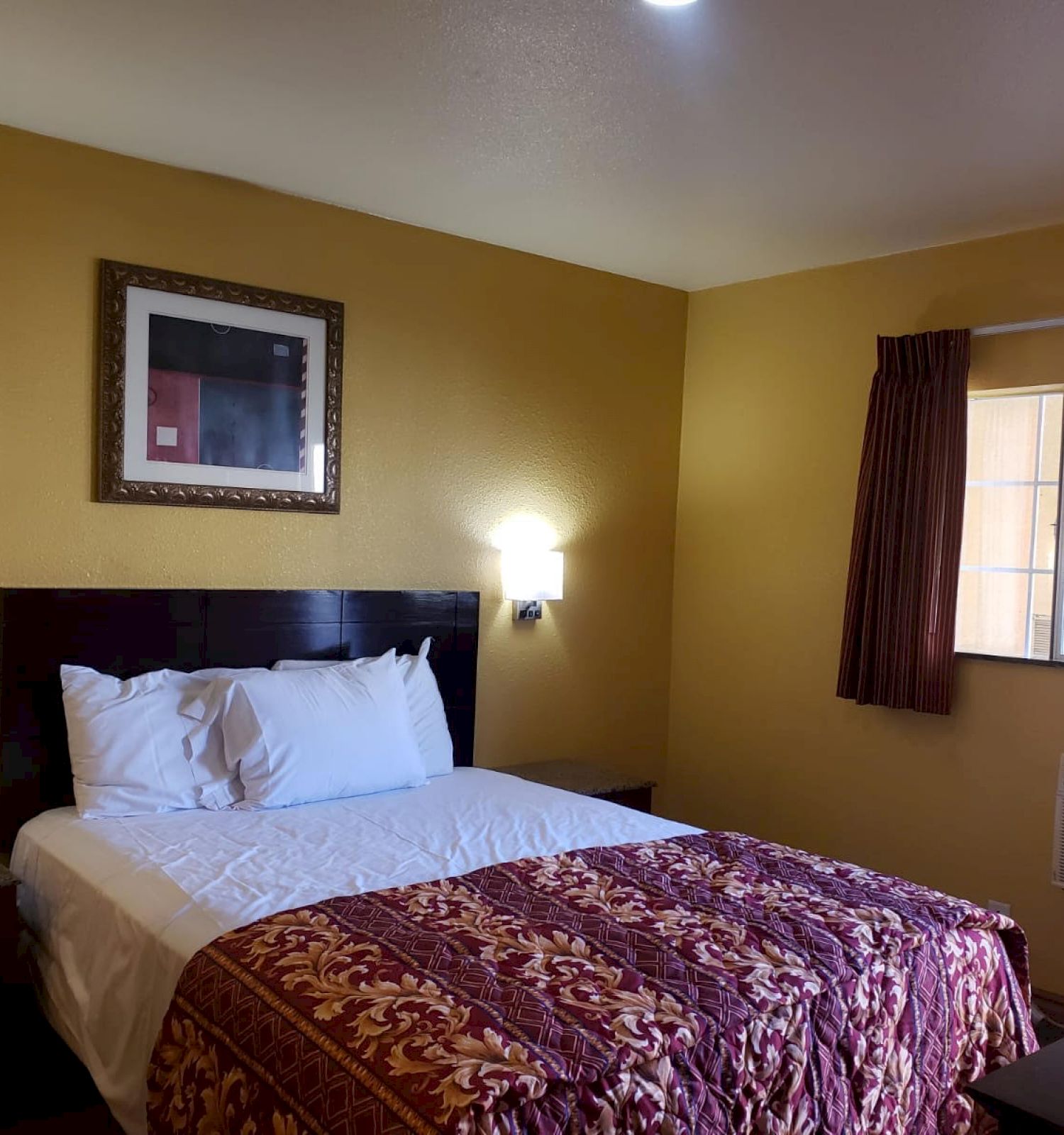 A hotel room with a bed, framed art, two wall lights, a desk, chair, window with curtains, and an air conditioning unit.