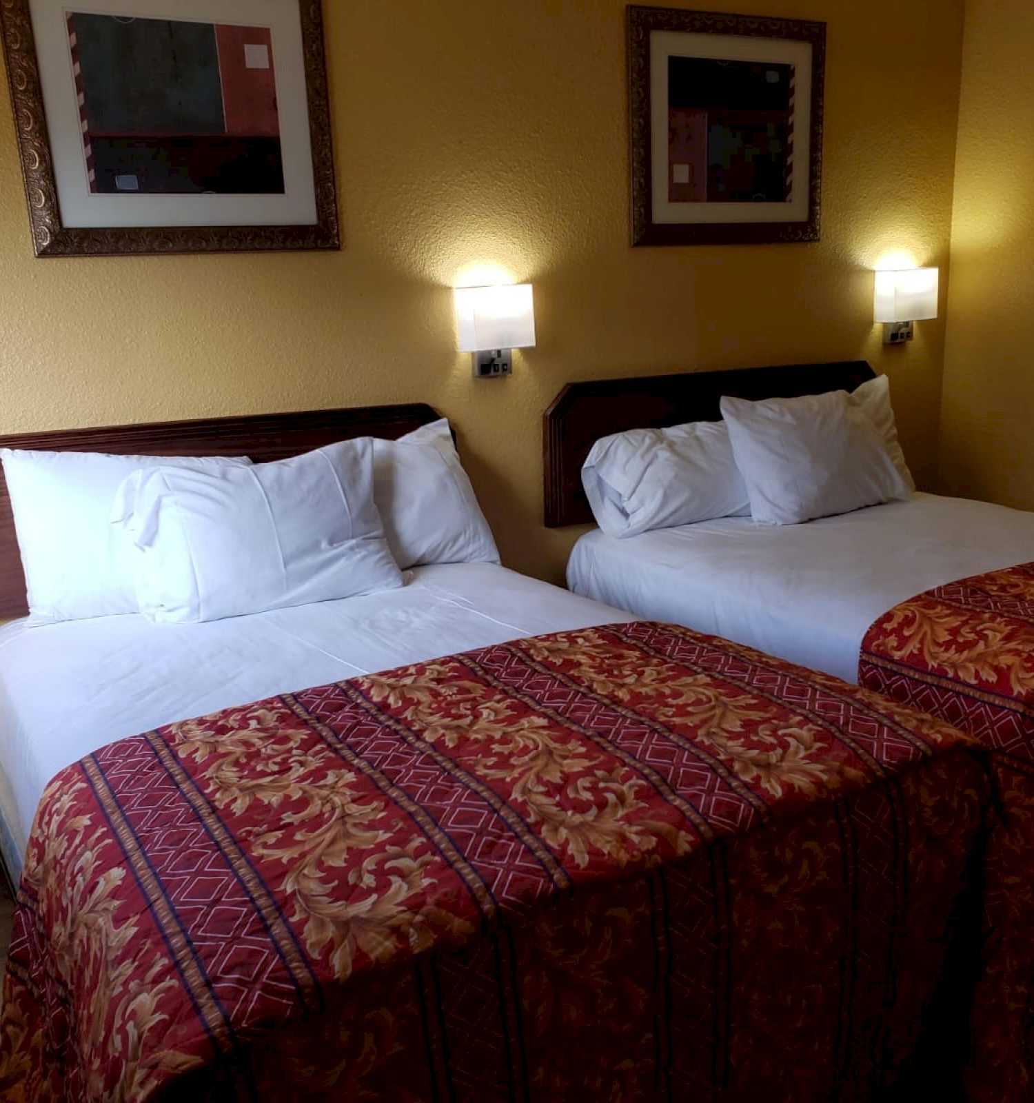 The image shows a hotel room with two neatly made double beds, wall lamps, framed artwork above the beds, and a warm yellowish wall color.