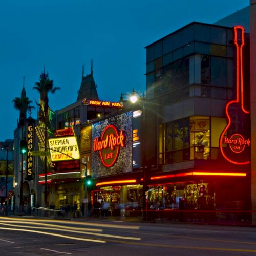 A bustling street at dusk with prominent Hard Rock Cafe sign, neon lights, and various storefronts under a darkening sky.